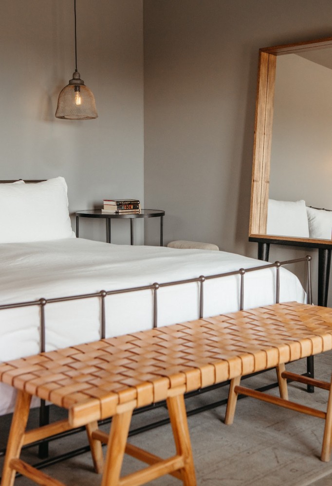 A neatly made bed with white linens and a wooden bench at the foot, with a nightstand and pendant light in the background.