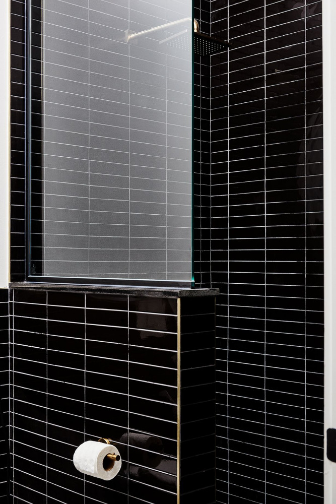 A black tiled bathroom with a glass shower door and a roll of toilet paper on the wall.