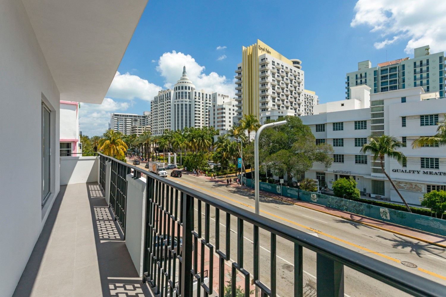 A balcony with a black railing overlooking a city street with palm trees and tall buildings.