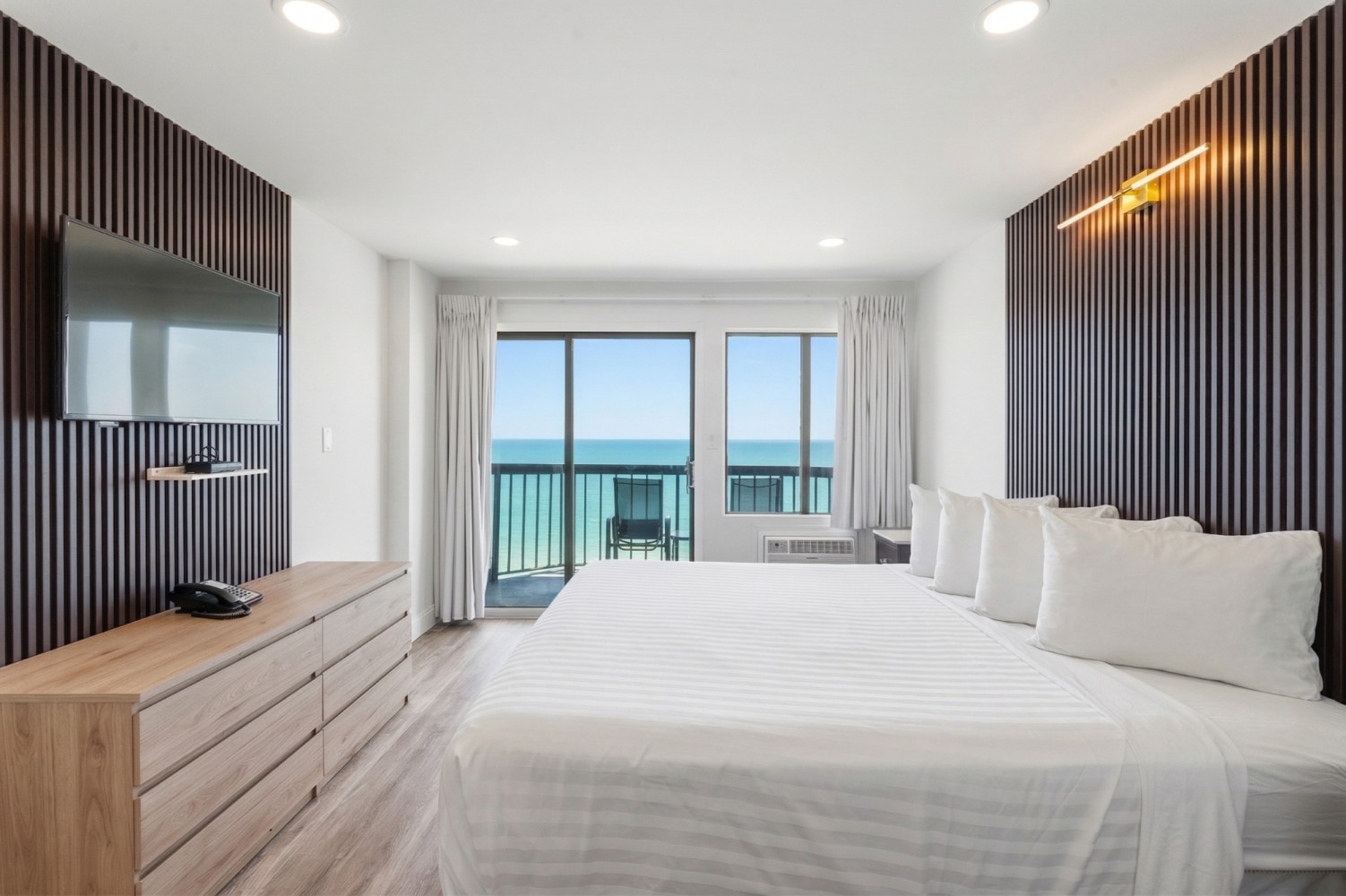 A wide view of a hotel room with a large bed, dresser, TV, and sliding glass doors leading to a balcony with an ocean view.