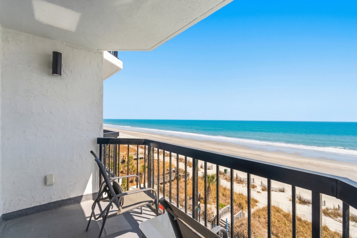 A balcony with a black railing and two chairs overlooking a beach and ocean.