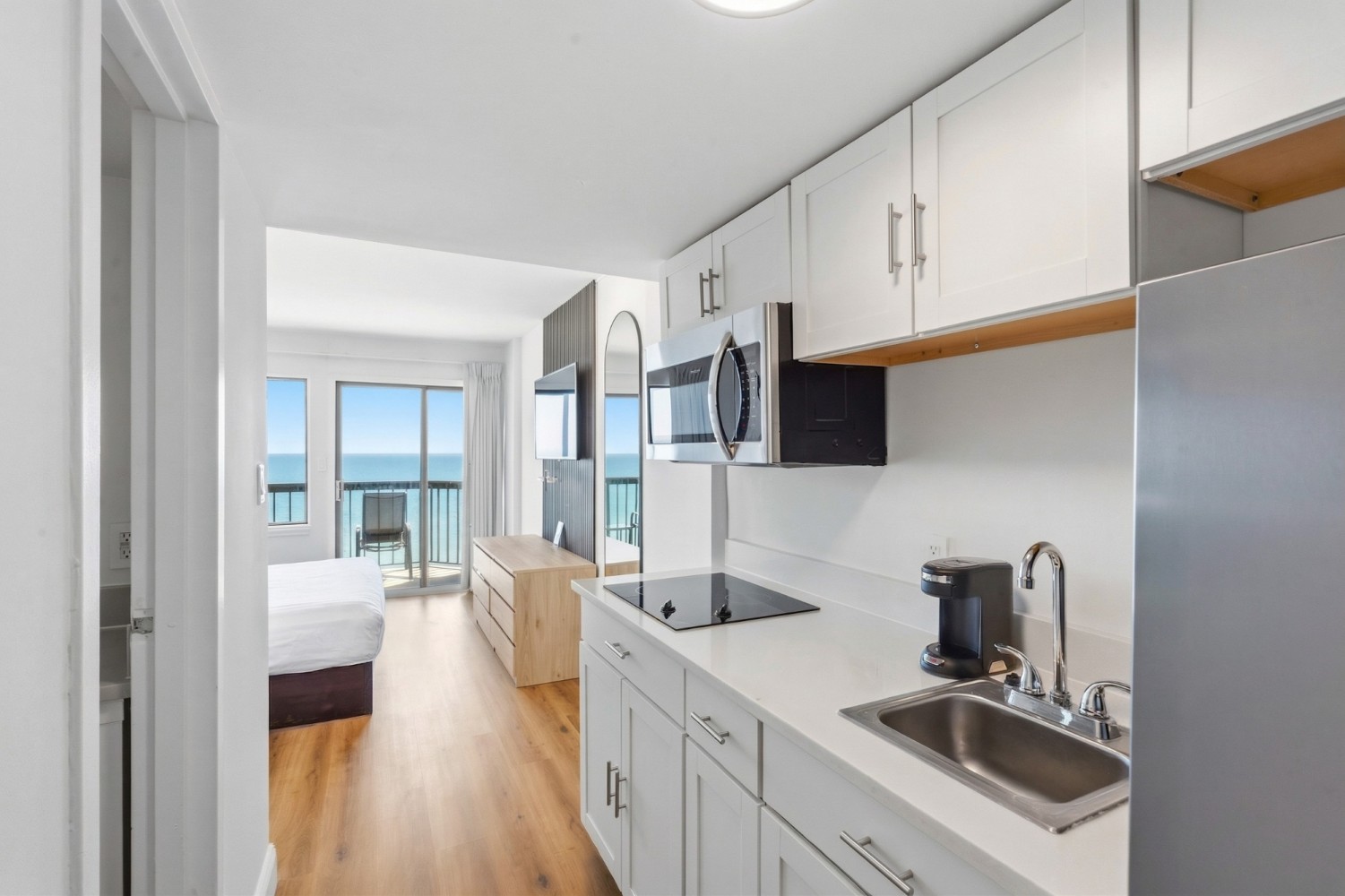 A compact kitchenette with white cabinets and stainless steel appliances, overlooking a bedroom with a bed and ocean view.