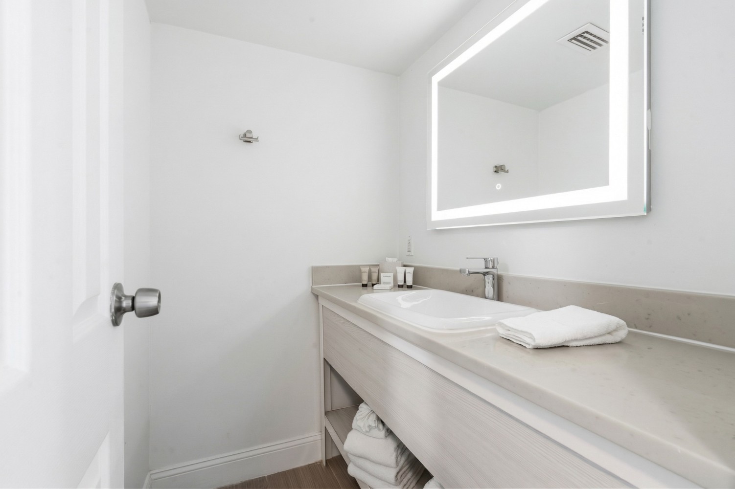 A clean and modern bathroom sink area with a lighted mirror, white towels, and wooden cabinetry.