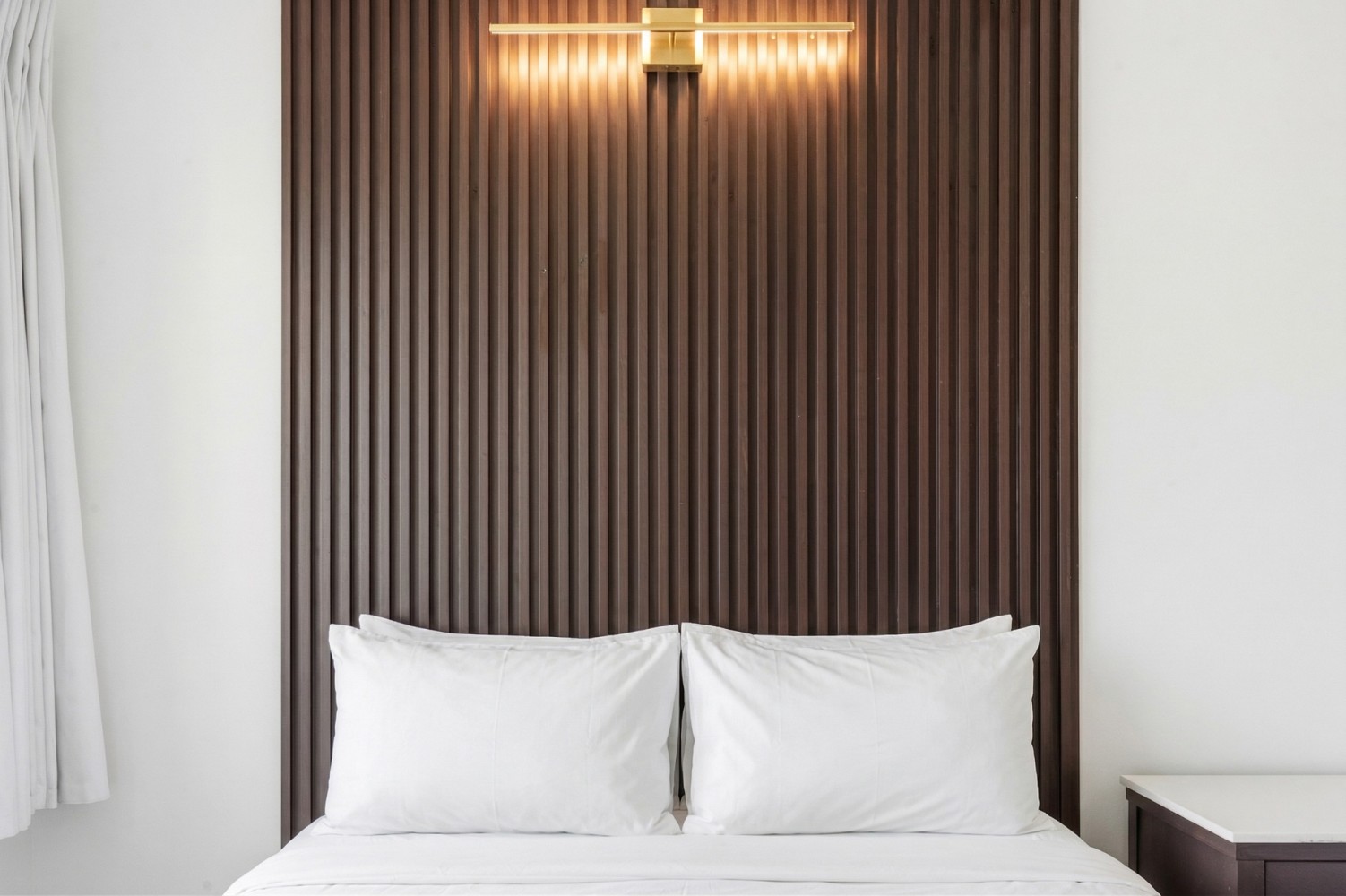 A neatly made bed with white pillows and bedding in front of a wood-paneled headboard with a wall light above it.