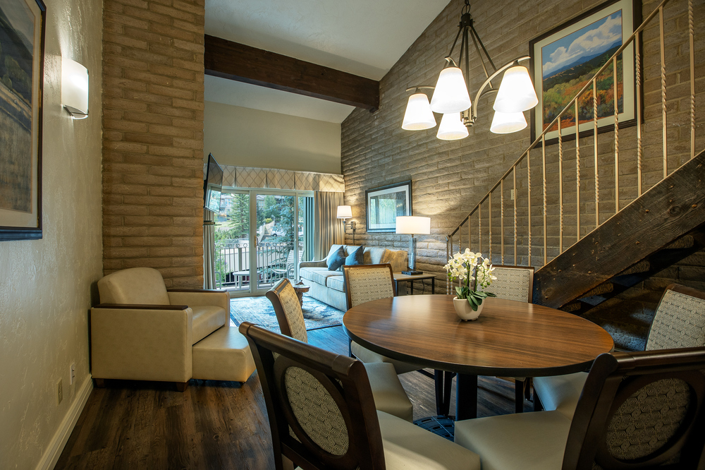 A wide view of a dining area with a round table and chairs, a staircase to the right, and a living room in the background.