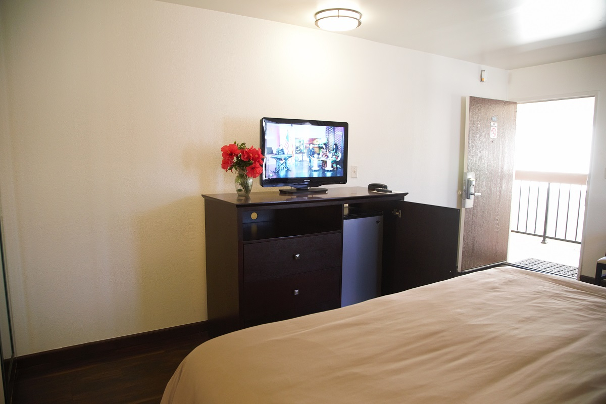 A hotel room with a bed, TV, mini-fridge, and flowers on a desk