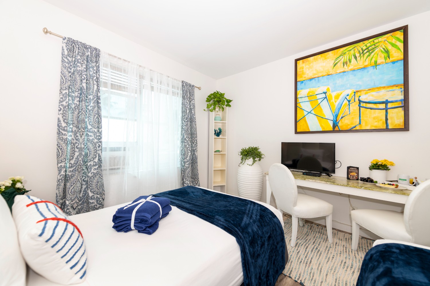 A bright and clean guest room with a bed, desk, TV, and artwork.