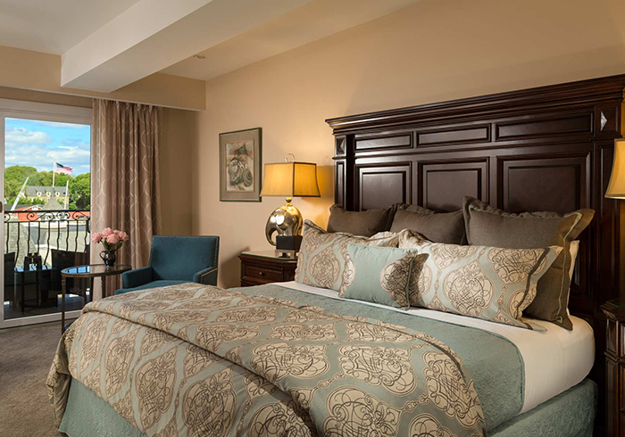 A wide-angle view of a hotel room with a large bed, dark wood headboard, and a window with a balcony overlooking the outdoors.