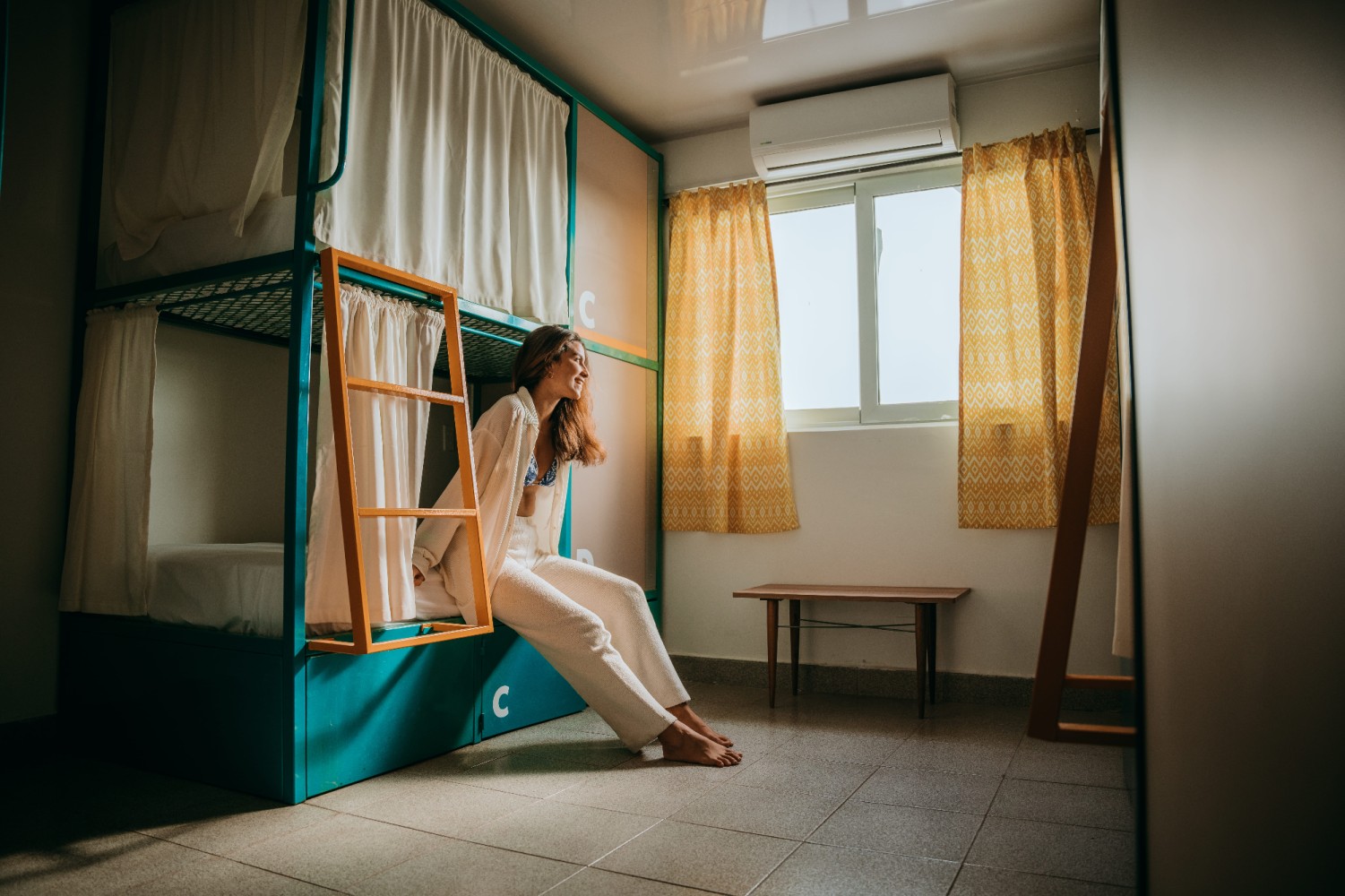 A woman sitting on the bottom bunk of a teal bunk bed in a small room with a window and yellow curtains.